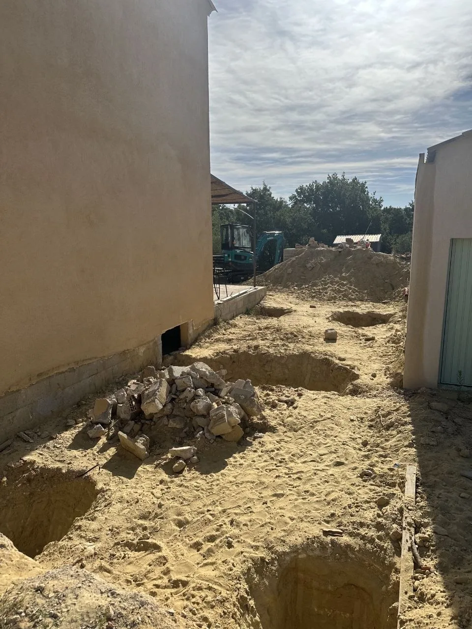 Photo de chantier : fouilles de fondation creusées le long des murs après décaissement, mini-pelle en arrière-plan